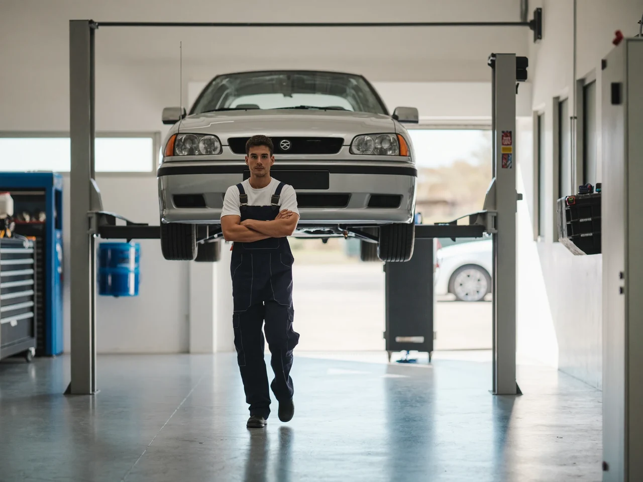 Ilustrace světlého dílenského stání s vozem na dvousloupovém zvedáku a mechanikem v overalu